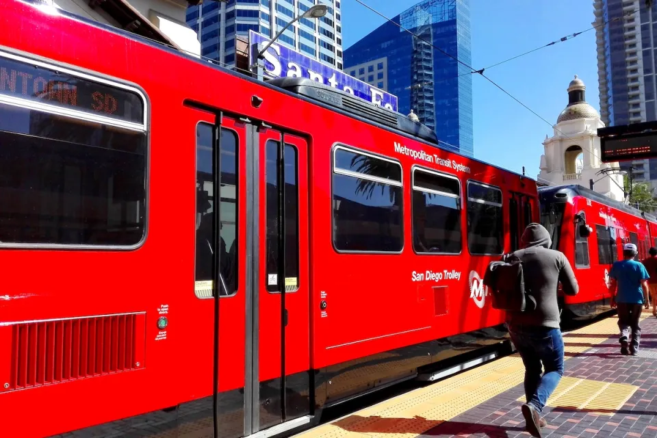 Red San Diego Trolley light rail train at station with passengers walking on platform and Spanish-style dome building in background.